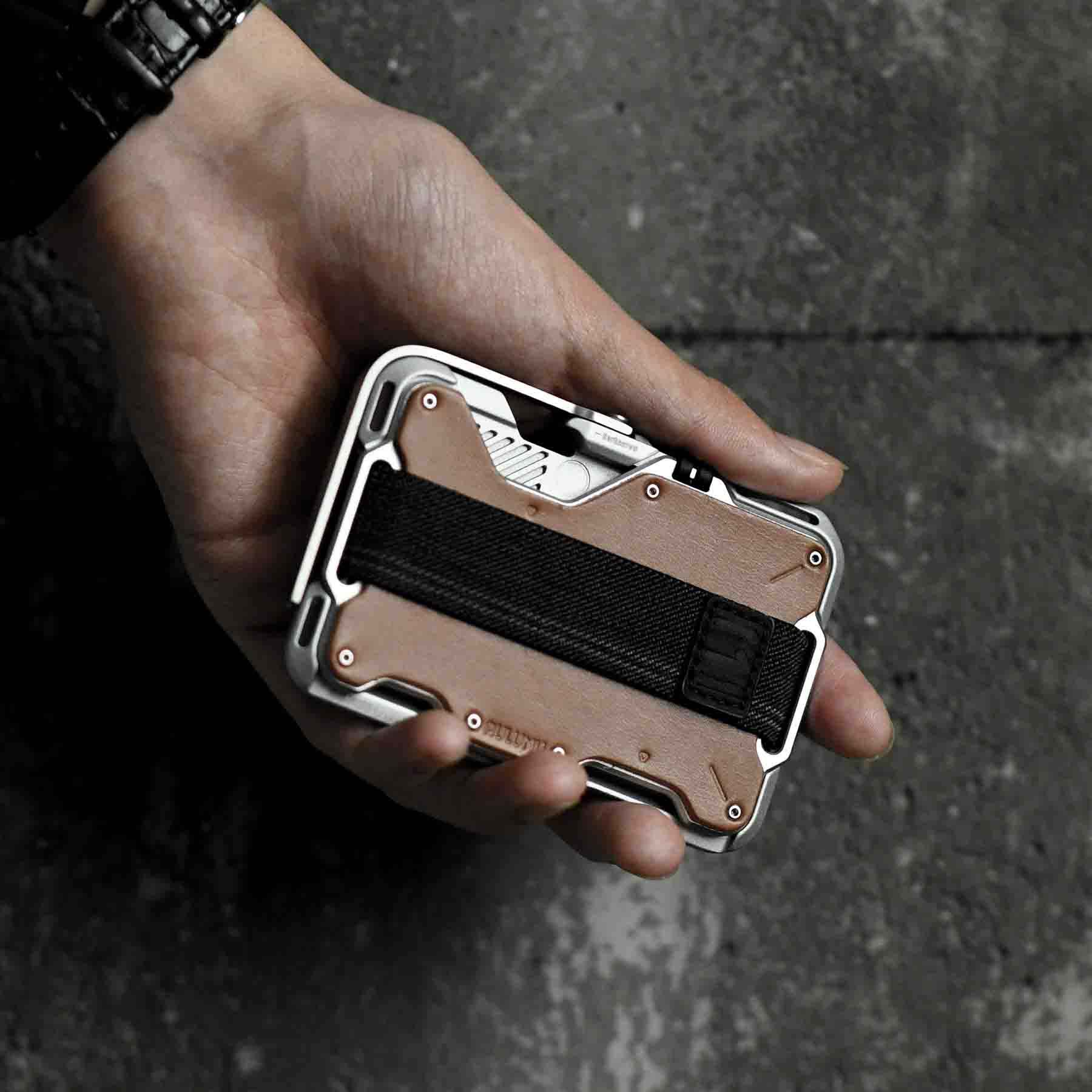 Hand holding a tactical wallet with a metallic frame and black strap on a dark background