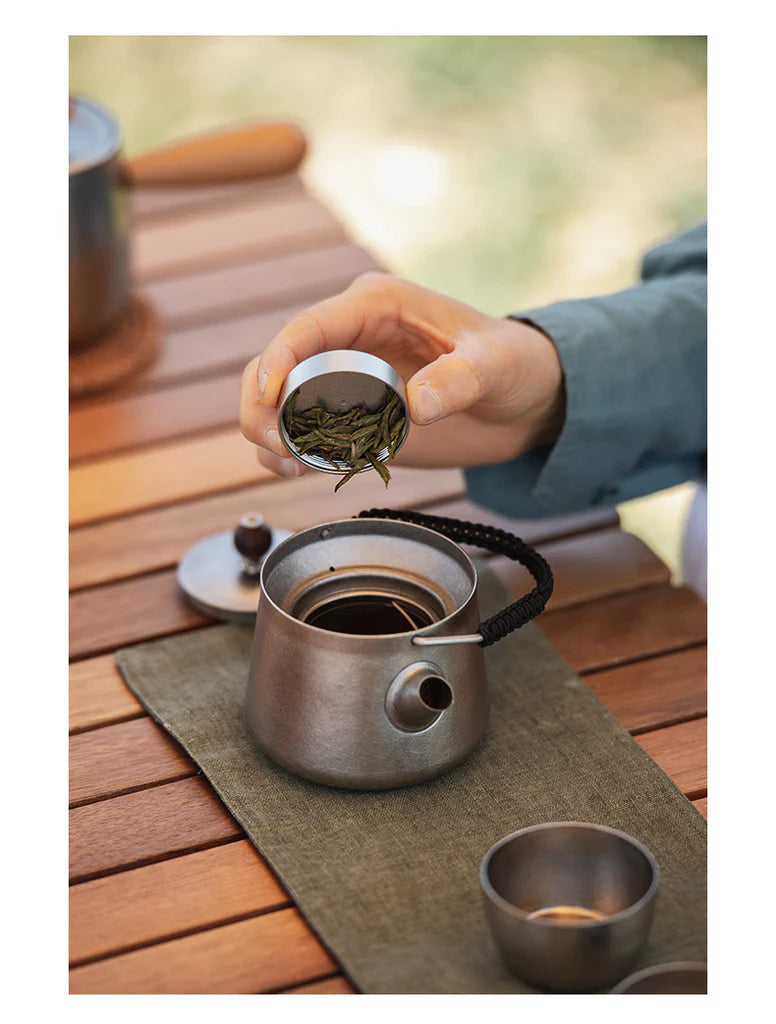 Person preparing tea by adding tea leaves to a teapot on a wooden table.
